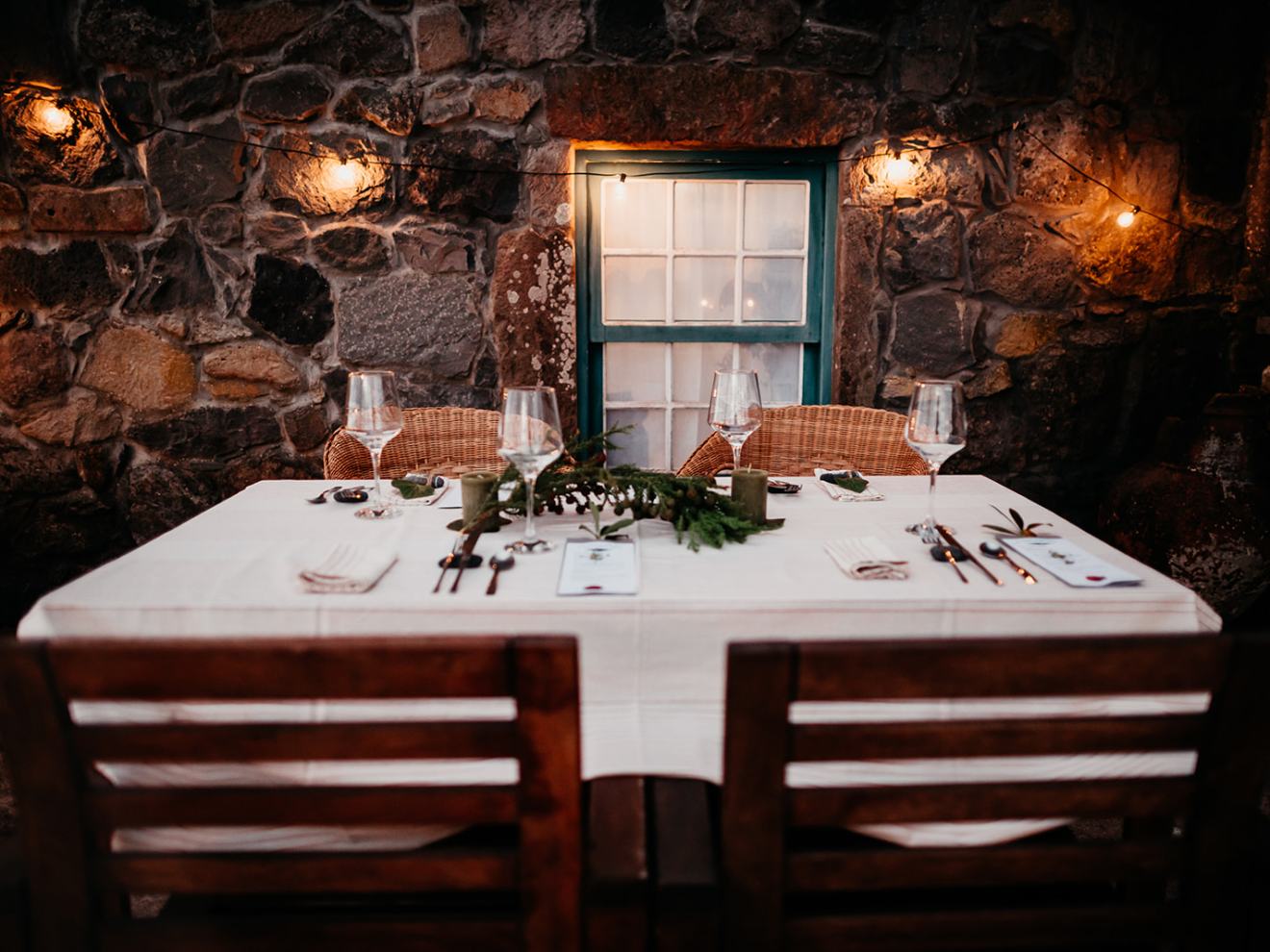 Rustic dining table set for four with bright string lights and a stone wall backdrop.