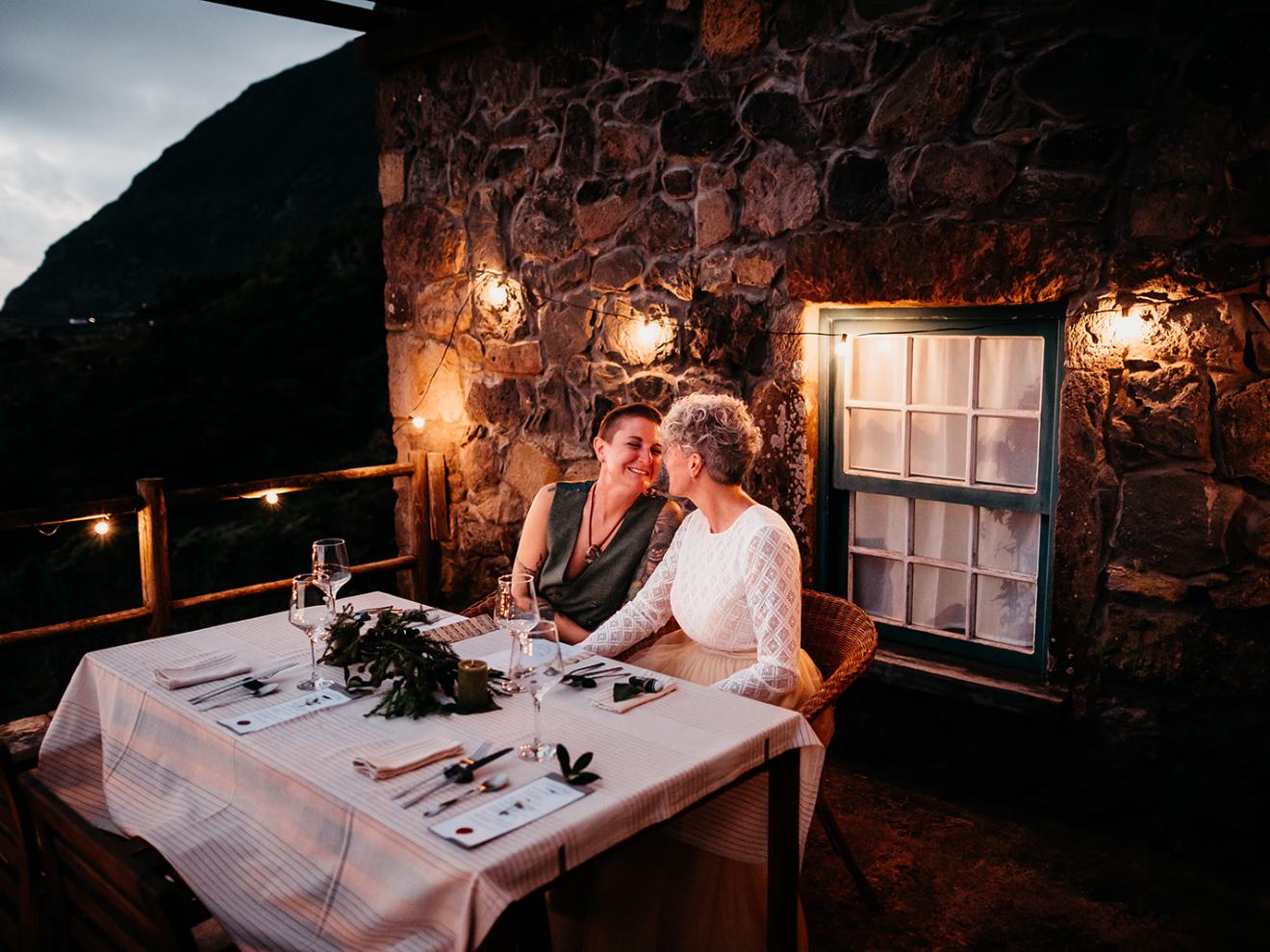 Two women smiling at a candlelit dinner table against a stone wall backdrop.