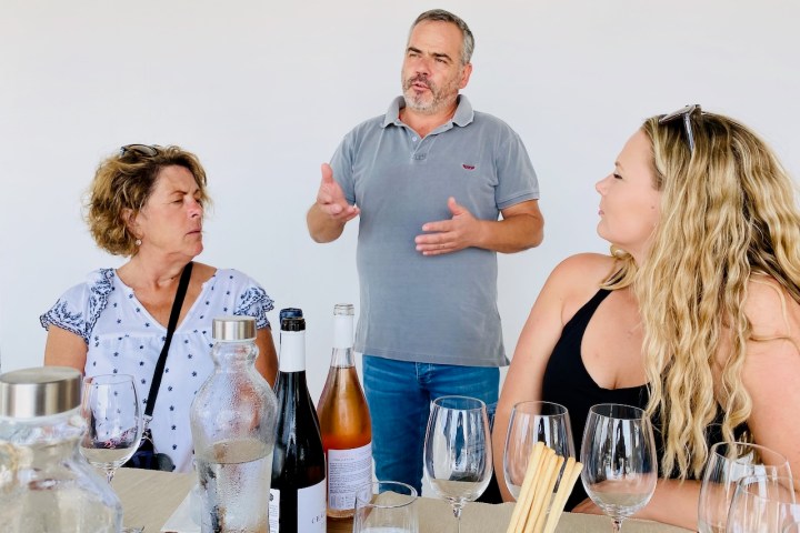Man talking to two women at a table with wine bottles.