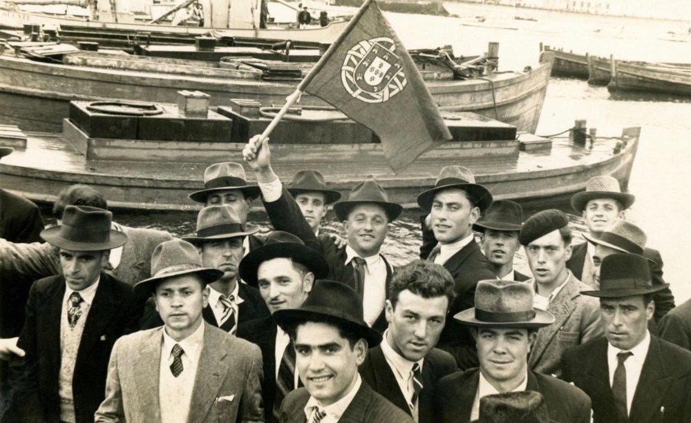 Vintage photo of men in hats holding a Portuguese flag near docked boats.