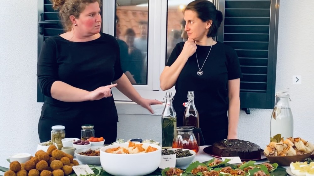 Two women in black shirts gesture over a table of assorted foods and drinks.