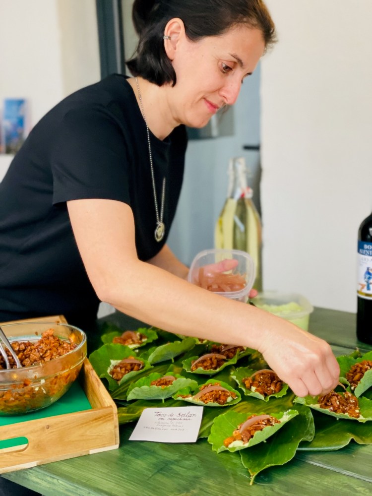 Person prepares green leaf-wrapped food on a table; bowl of filling and bottles nearby.