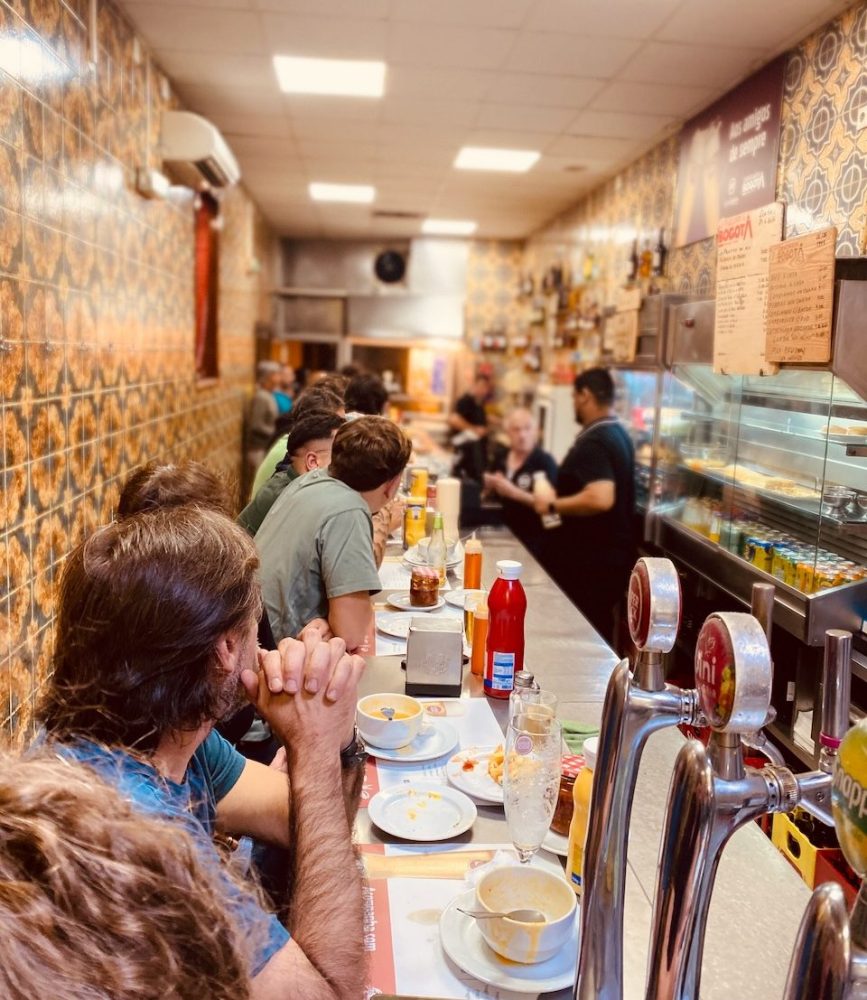 People seated at a narrow cafe counter with a barista serving drinks.