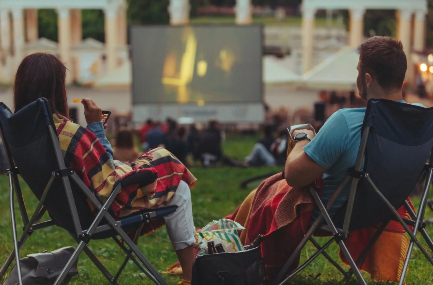 Two people sit in chairs with blankets, watching an outdoor movie on a large screen.