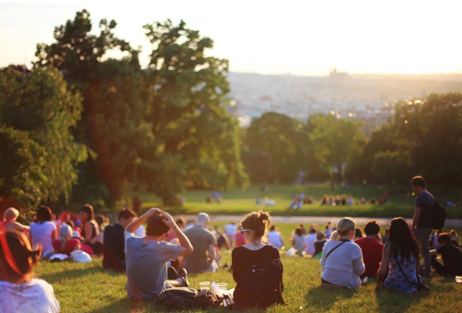 People sitting on grassy hillside in a park during sunset with city view in background.