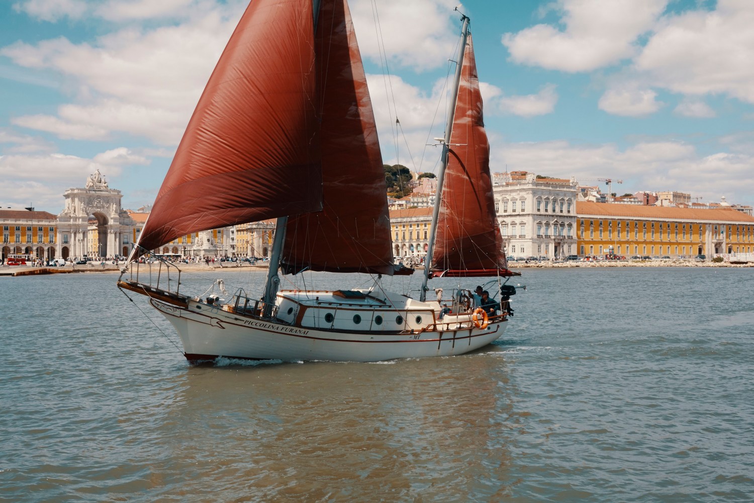 Sailboat with red sails on a river near historic buildings under a blue sky with clouds.