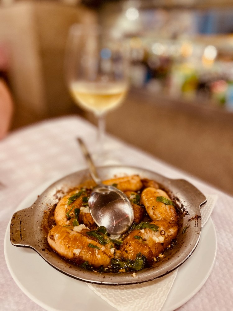 A dish of cooked shrimp with herbs and a spoon, next to a glass of white wine.
