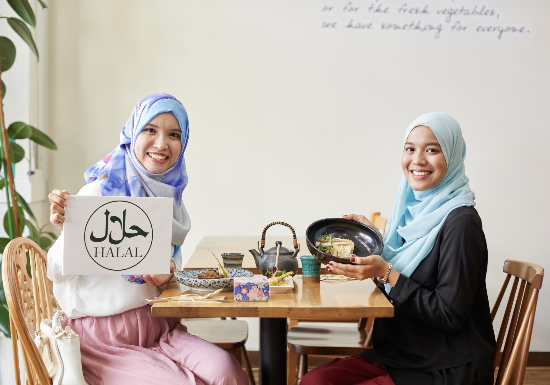 Two women at a table showcasing halal food and a halal sign.